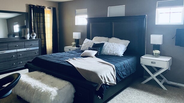 Quarto com cabeceira em madeira escura, uma cama grande com roupa de cama branca e azul escura, cómoda branca, mesa de cabeceira branca com uma luminária e uma janela com persianas.