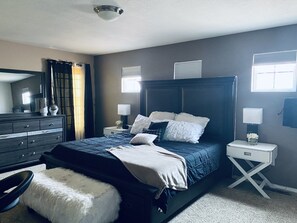 A bedroom with a dark wood headboard, a large bed with white and dark blue bedding, a white dresser, a white nightstand with a lamp, and a window with blinds.