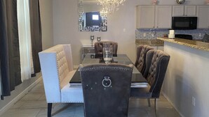 Uma sala de jantar com um candelabro, uma televisão e uma cozinha ao fundo.