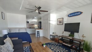 Een woonkamer met een bank, salontafel en televisie. Achtergrond is een keuken en een bord met daarop 'Beach House'.