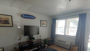 Een woonkamer met een televisie, een bordje met de tekst 'Beach House', een kamerkleed met een patroon en een raam met gordijnen.