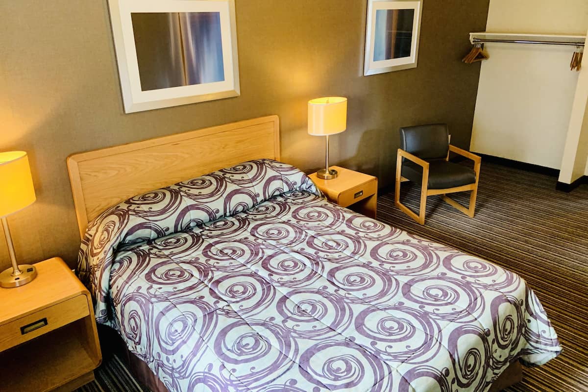 A hotel room with a bed, a nightstand with a lamp, a chair, and two framed pictures on the wall.
