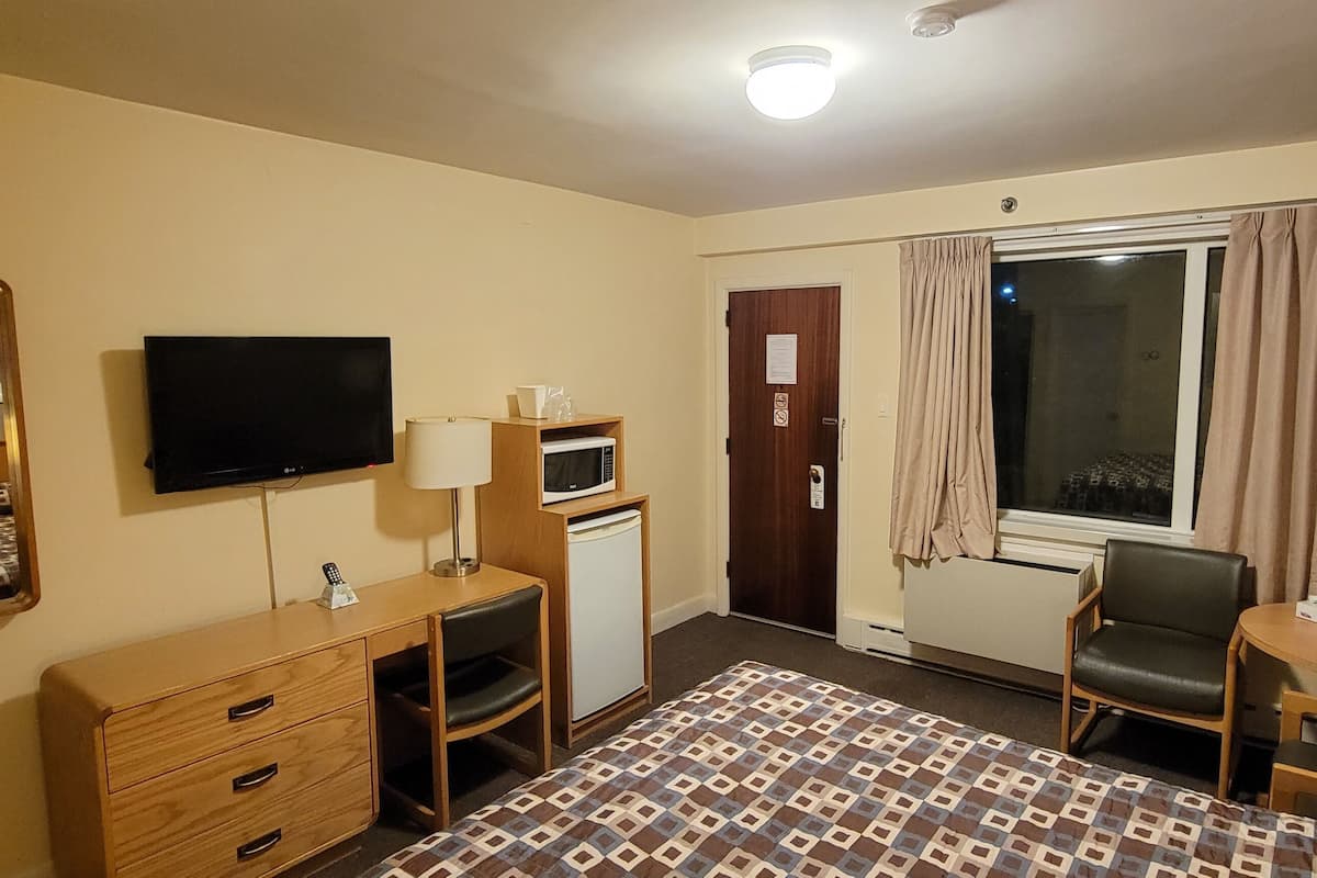 A hotel room with a bed, a desk, a television, a microwave, a small refrigerator, a chair, and a window with curtains.