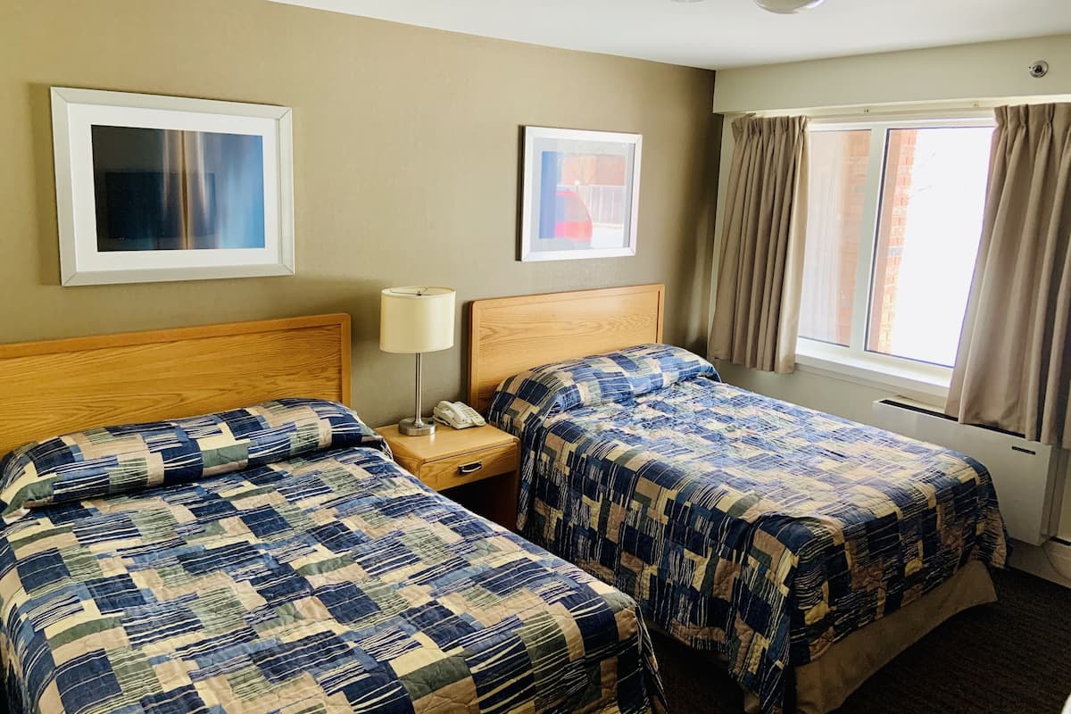 A hotel room with two beds, a bedside table with a lamp, and a window with curtains.