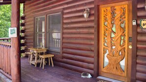 Eine Holzhütte mit einem großen Baumdesign an der Haustür, einer Veranda mit Tisch und Stühlen und einem Schild, das daraufhinweist, dass die Hütte vermietet wird.