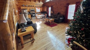 Ein gemütliches Blockhausschlafzimmer mit Holzboden, einem Essbereich, einem Wohnzimmer mit Sofa und Couchtisch sowie einer Küche mit Holzausstattung und einem festlich geschmückten Weihnachtsbaum.