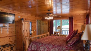 Ein Schlafzimmer einer Holzhütte mit einem Bett, einem Deckenventilator, einem an der Wand montierten Fernseher und einem Fenster mit Vorhängen.
