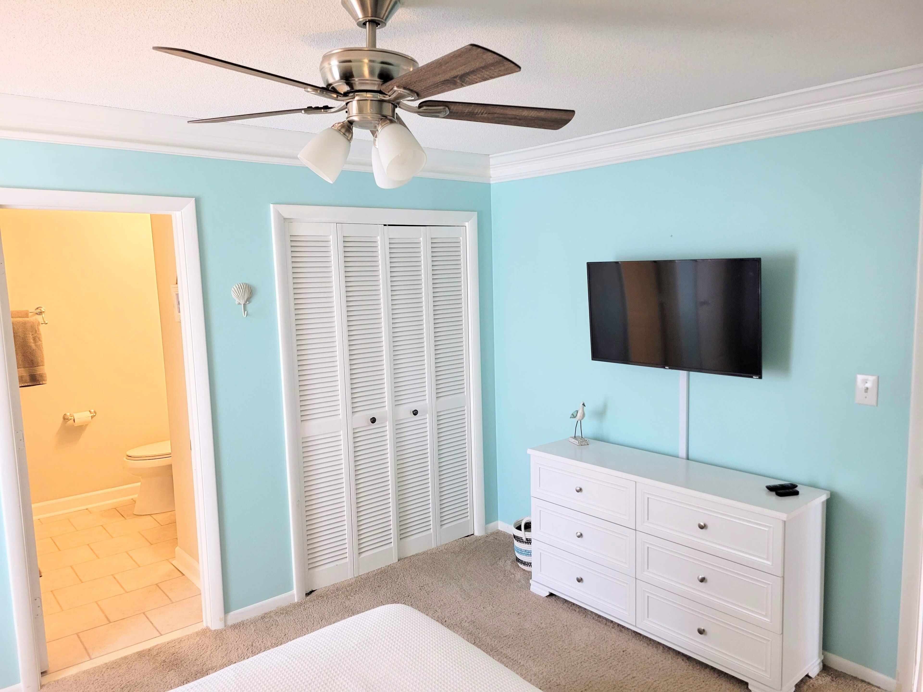 A bedroom with a ceiling fan, a white dresser, a television on the wall, and a closet with white shutters.
