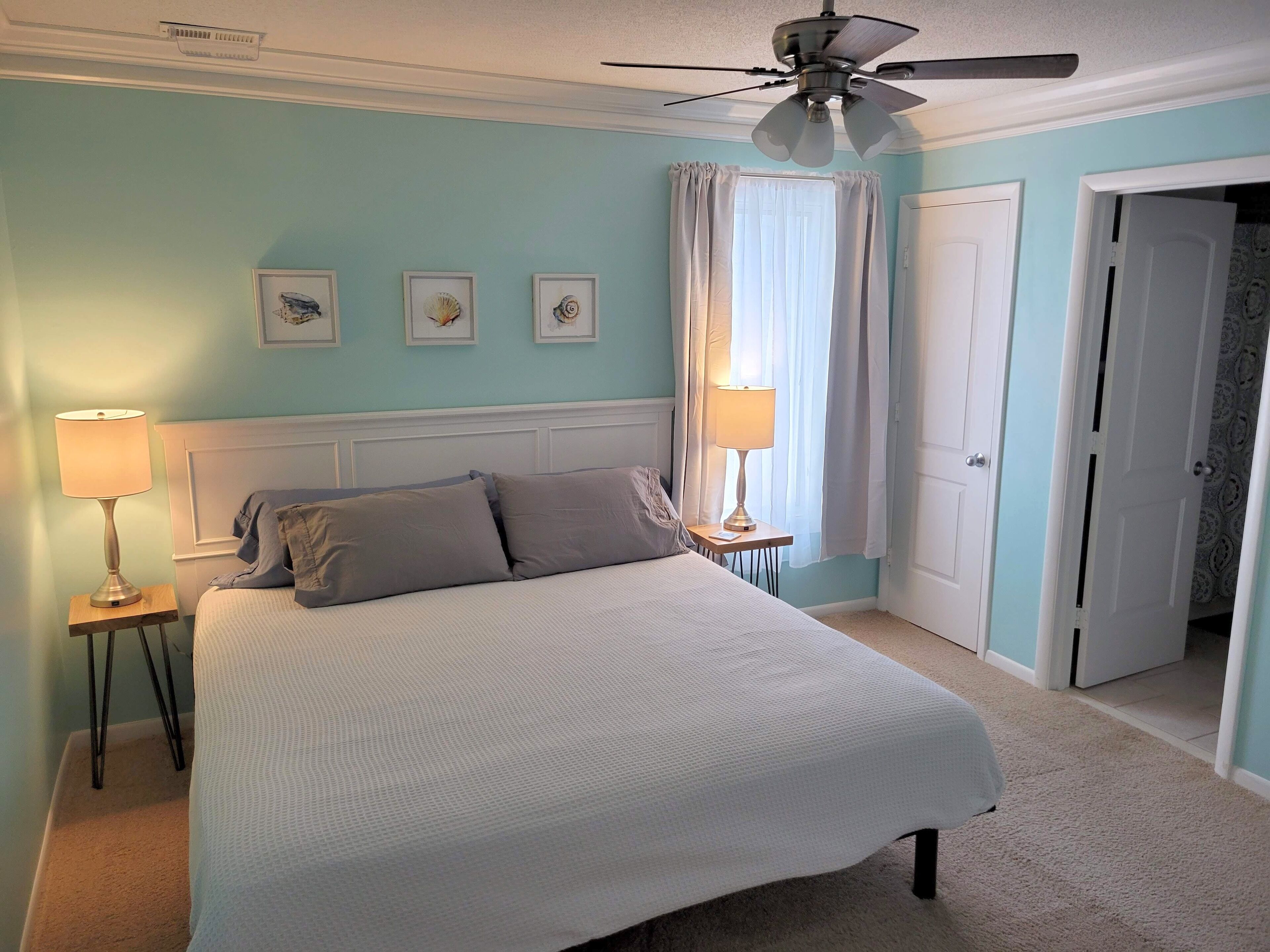 A bedroom with a bed, two bedside tables with lamps, a ceiling fan, and a door leading to another room.