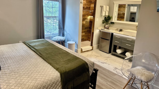 Een slaapkamer met een bed, een transparante stoel, een kleine badkamer met een spiegel en een douche.