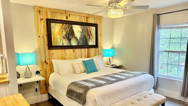 A bedroom with a bed, bedside lamps, a bench, a ceiling fan, a chandelier, a painting, and a window. - Suite 201 "The Golden Eagle" (Custer)