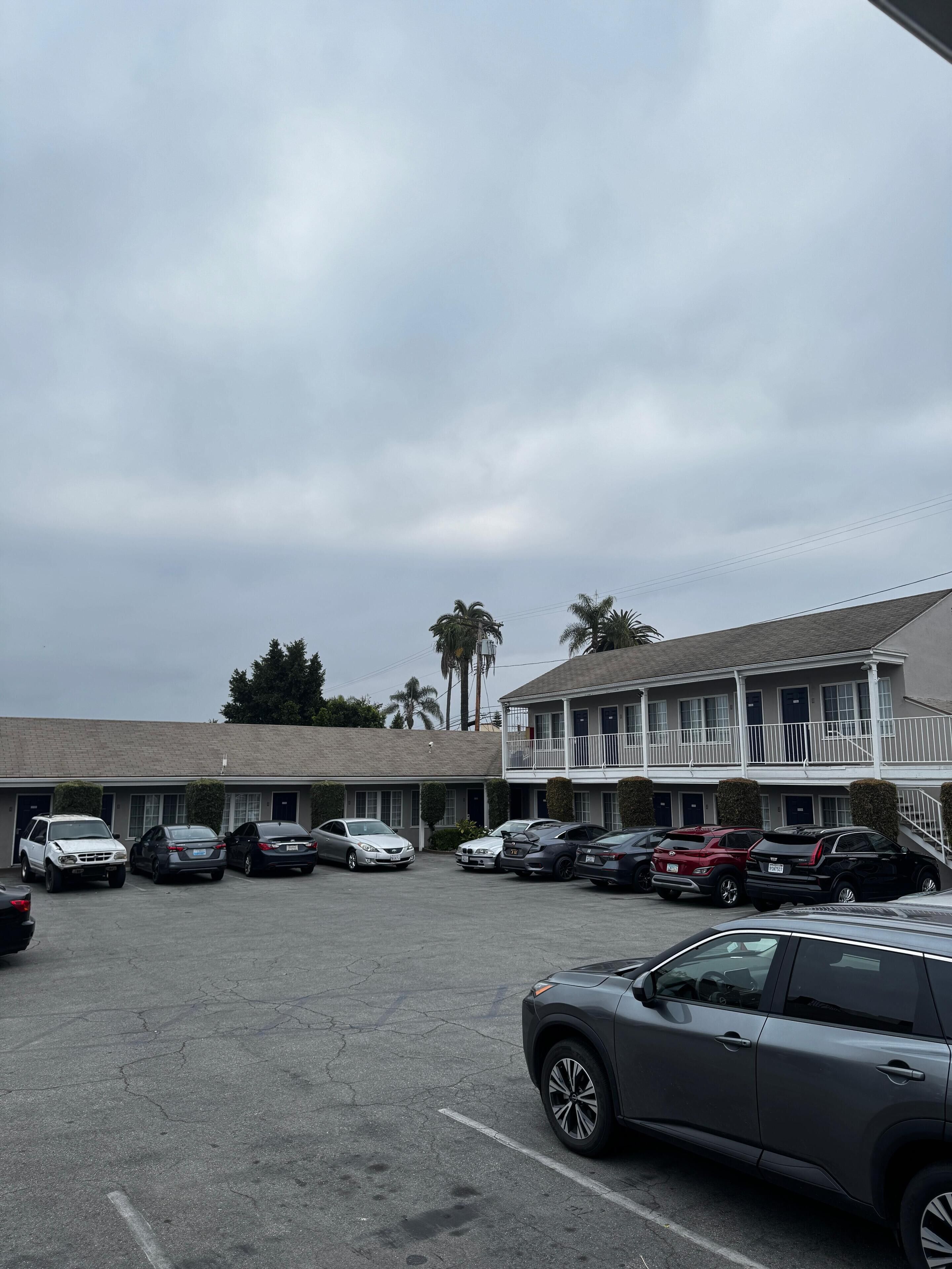 Un estacionamiento con varios automóviles, un edificio de motel con balcón de dos pisos y palmeras al fondo.