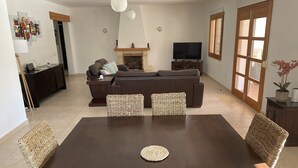 Un salon moderne avec une table à manger en bois foncé, deux petites tables d’appoint en osier, un canapé et un téléviseur.