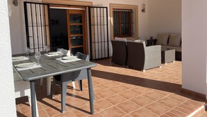 Une terrasse avec une table à manger pour quatre personnes, deux canapés en osier et une fenêtre ornée d’une grille métallique.