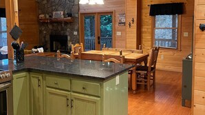 A cabin interior with a kitchen featuring a granite countertop, green cabinets, a stone accent wall, a fireplace, and a dining area with wooden furniture. - Spacious Blue Ridge Mountain Cabin. Great location & beautiful views!  (Morganton)