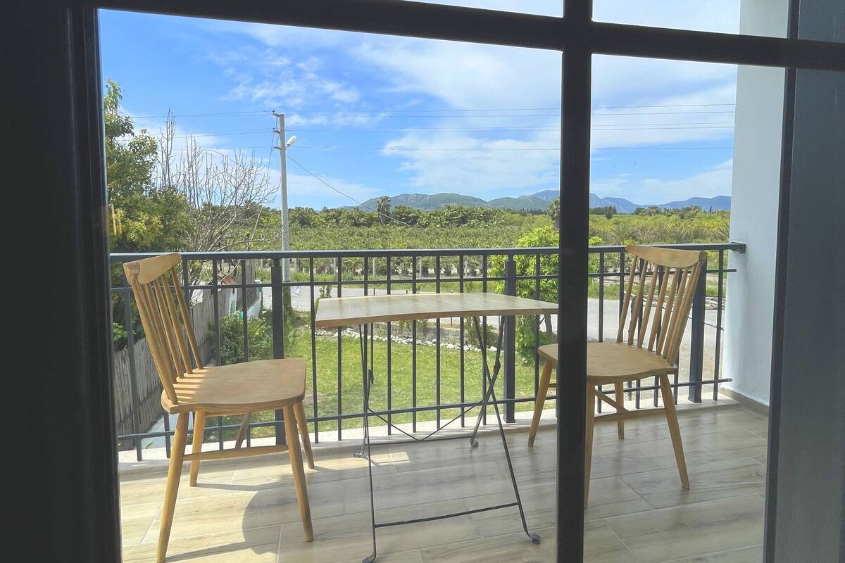 A balcony with a wooden table and chairs, a metal railing, and a view of a green landscape with trees and hills.