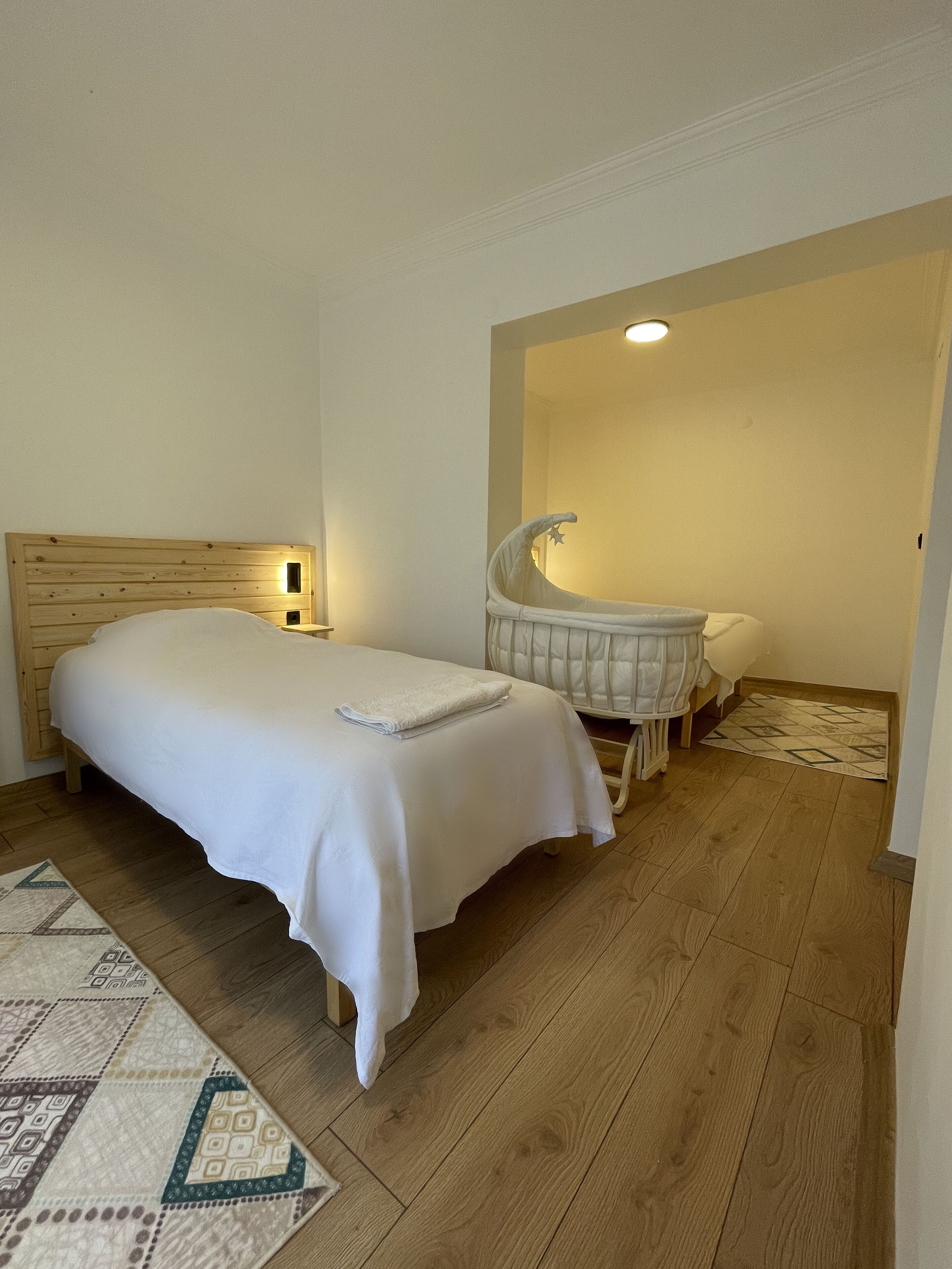 A bedroom with a wooden headboard, a white bed, a bedside lamp, a crib, and a patterned rug.