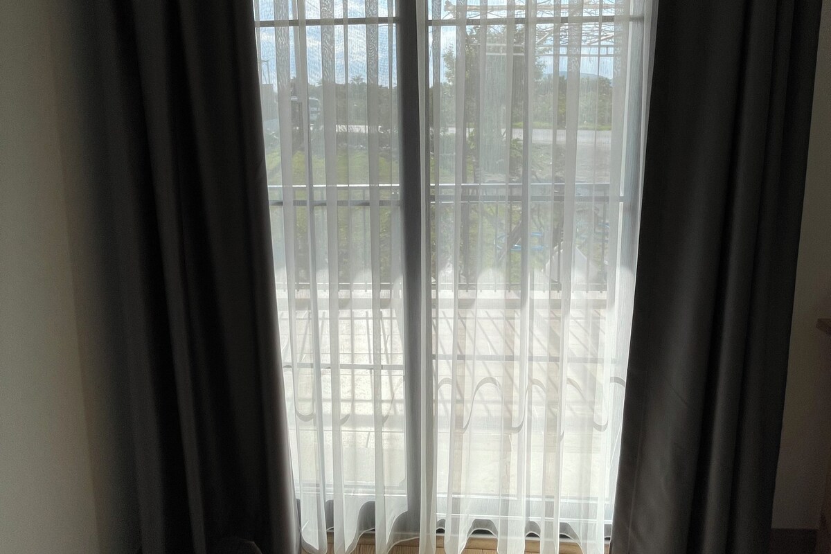 A room with a large window covered by sheer white curtains and dark drapes.