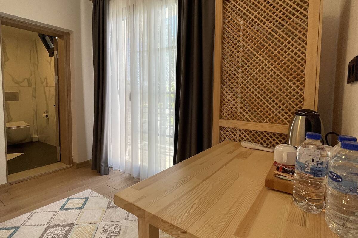 A room with a wooden table, a patterned rug, a window with curtains, a wooden partition, and a view of a bathroom.