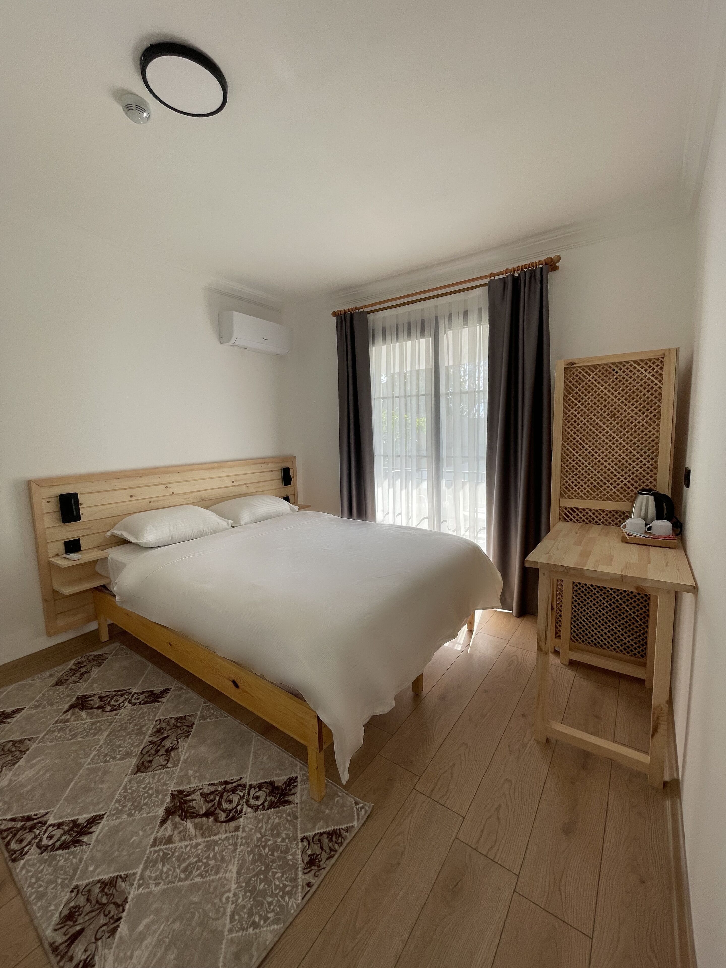 Un dormitorio con una cama de madera, una mesita de noche, un escritorio pequeño de madera, un tapete estampado y una ventana con cortinas.