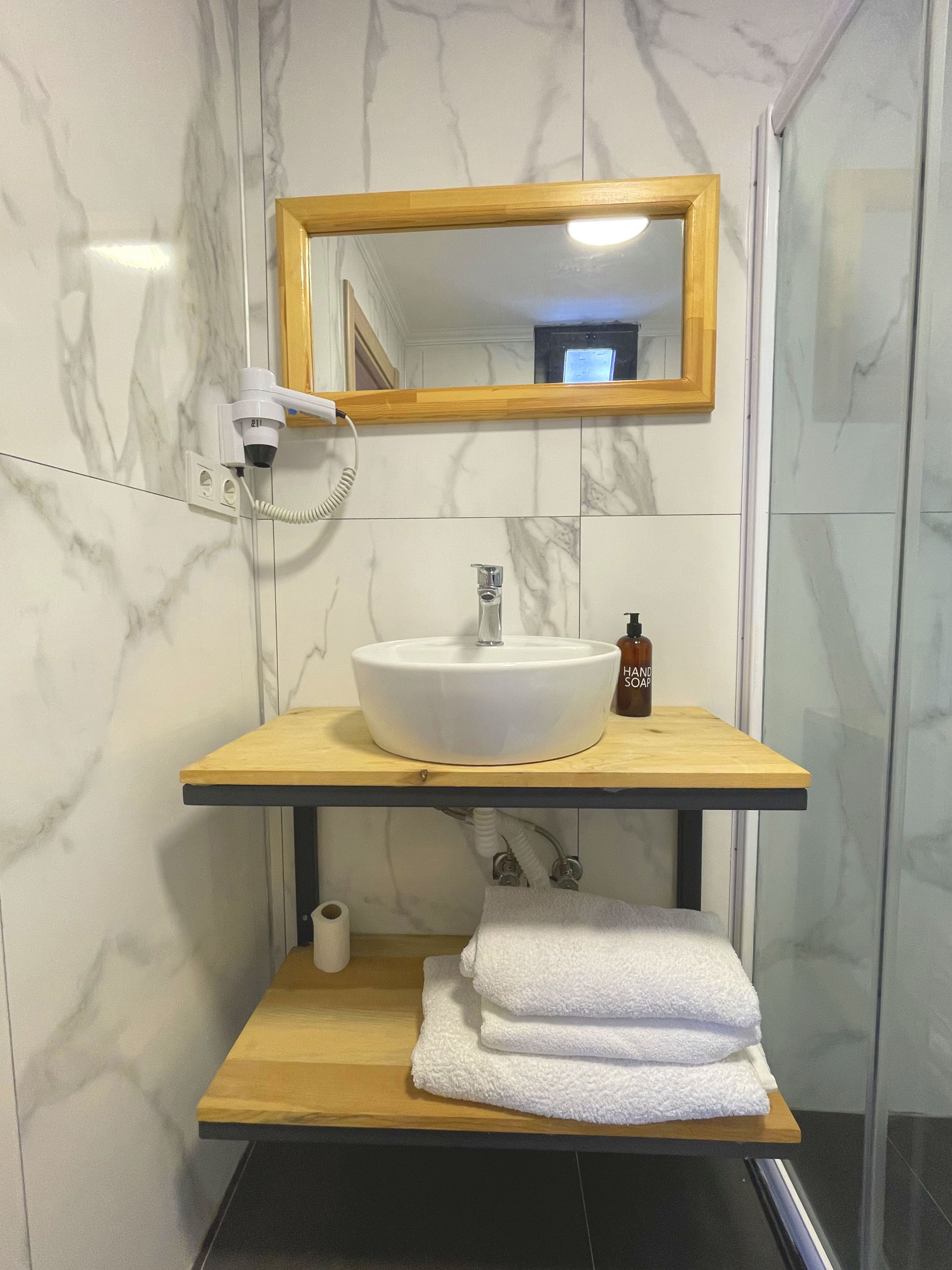 Un baño con pared de mármol, tocador de madera, lavabo blanco, espejo, dispensador de jabón de mano y una pila de toallas blancas.