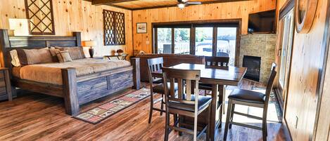 Interior de una cabaña de madera con una cama, una mesa de comedor, sillas, chimenea y un ventilador de techo.