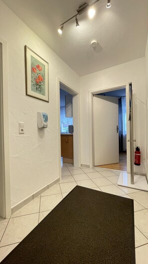 A hallway with white walls, a framed painting, a hand sanitizer dispenser, a fire extinguisher, and a tiled floor.
