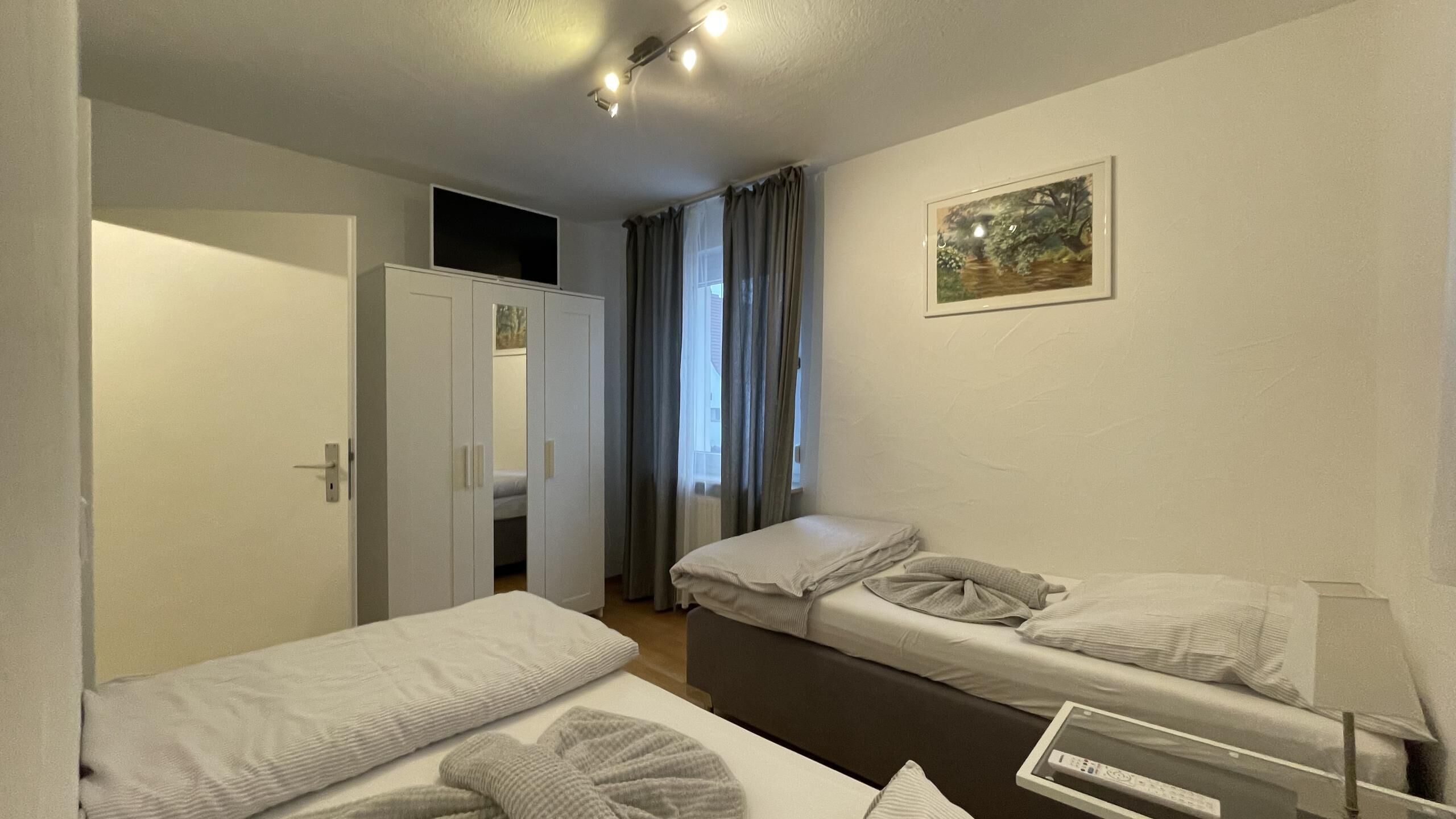 A small, well-lit room with two single beds, a wardrobe, a desk with a computer, and a window with curtains.