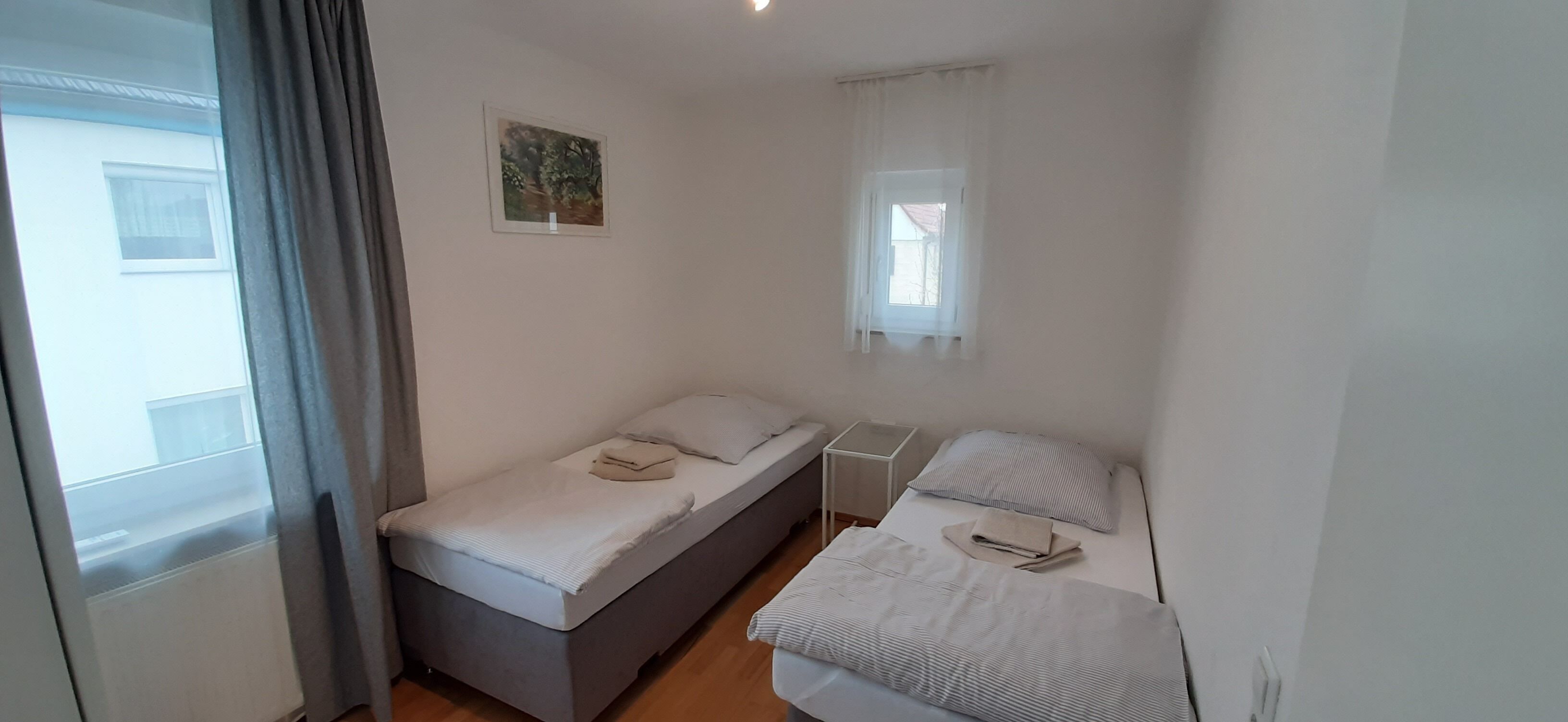 Ein kleines, sauberes Zimmer mit einem Einzelbett, einem Nachttisch und einem Fenster mit Vorhängen.