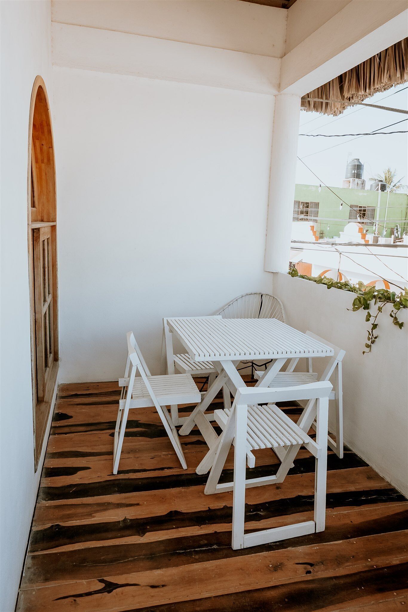 A small balcony with a white table and chairs, wooden flooring, and a view of buildings in the background.