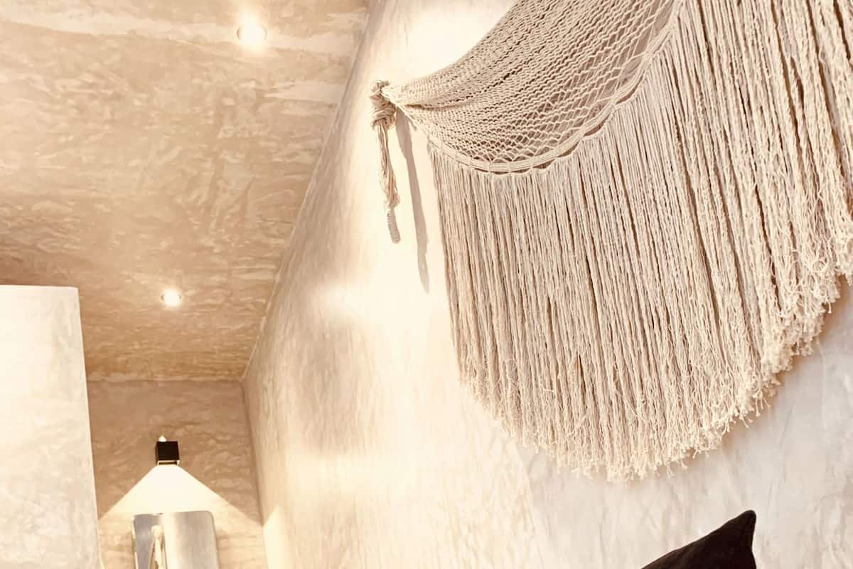 Une chambre à coucher avec un lit, des oreillers, une décoration murale en macramé, un miroir et un luminaire.