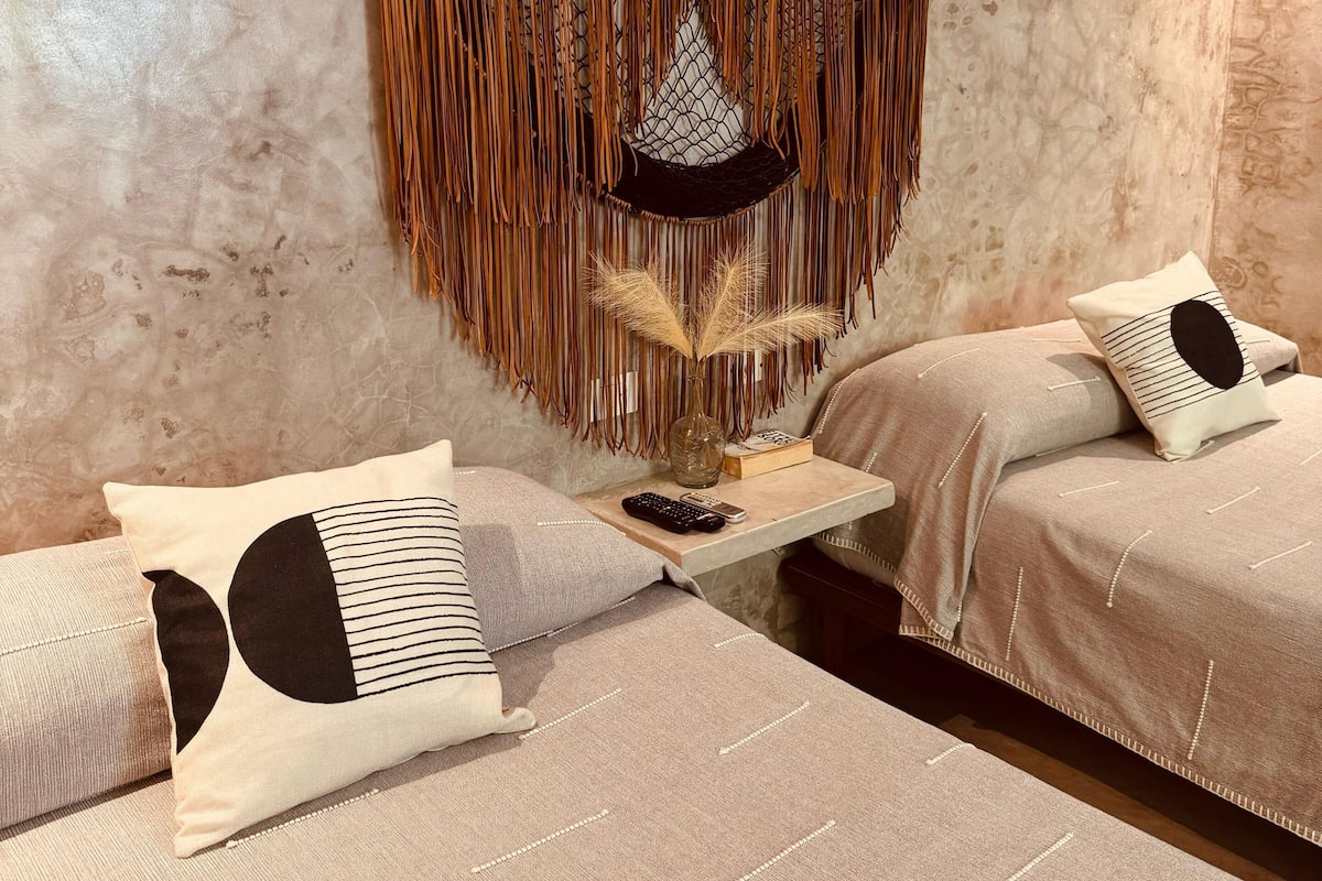 Une chambre avec deux lits, un luminaire suspendu en macramé et une petite table sur laquelle se trouvent un vase et des télécommandes.