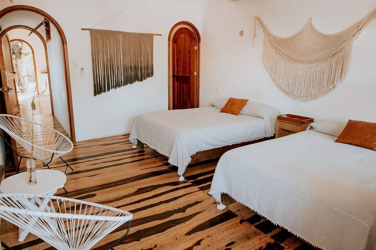 Une chambre avec deux lits, un plancher en bois, une chaise blanche et une décoration murale en macramé suspendue.