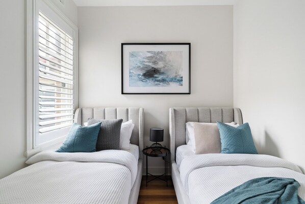 A bedroom with two beds, a framed abstract painting, a bedside lamp, and a window with blinds.