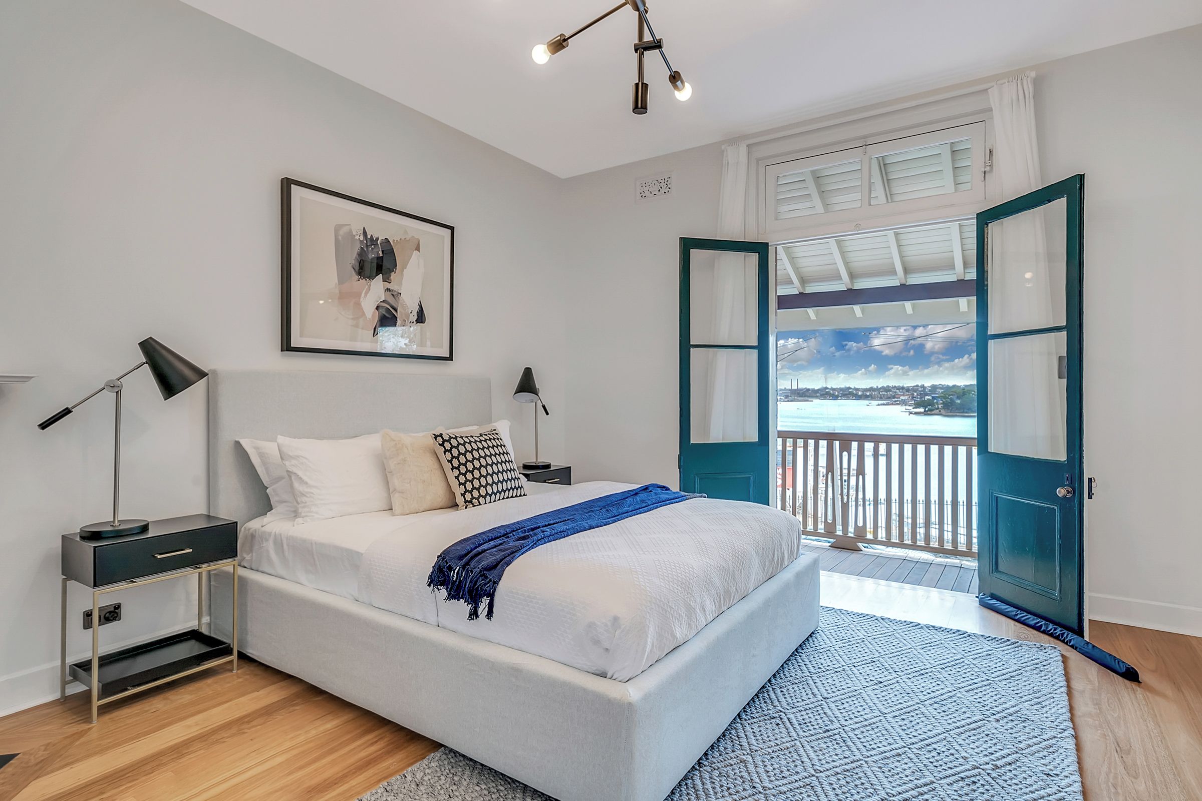 A bedroom with a bed, bedside tables, a lamp, a framed artwork, and a view of the sea through an open door.