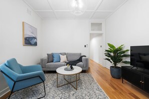 A modern living room with a grey sofa, a blue armchair, a round coffee table, a large potted plant, a black TV stand, and a painting on the wall. - Harbour View Terrace In The Heart Of The Rocks (Millers Point)