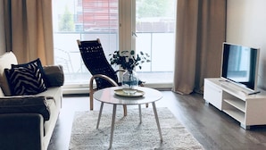A living room with a sofa, a chair, a coffee table, a TV, and a window with blinds. - Kotimaailma Pori Center (Pori)