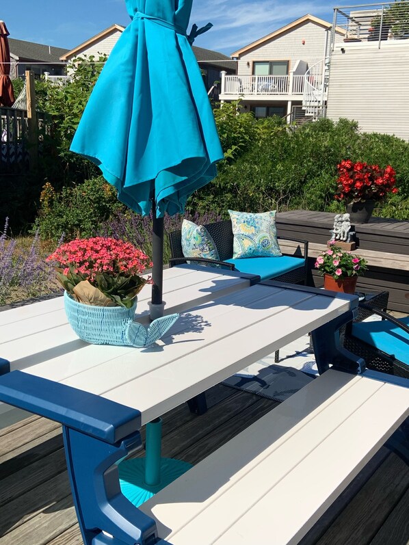 Un patio al aire libre con una mesa blanca y azul, un paraguas verde azulado y varias plantas en macetas.