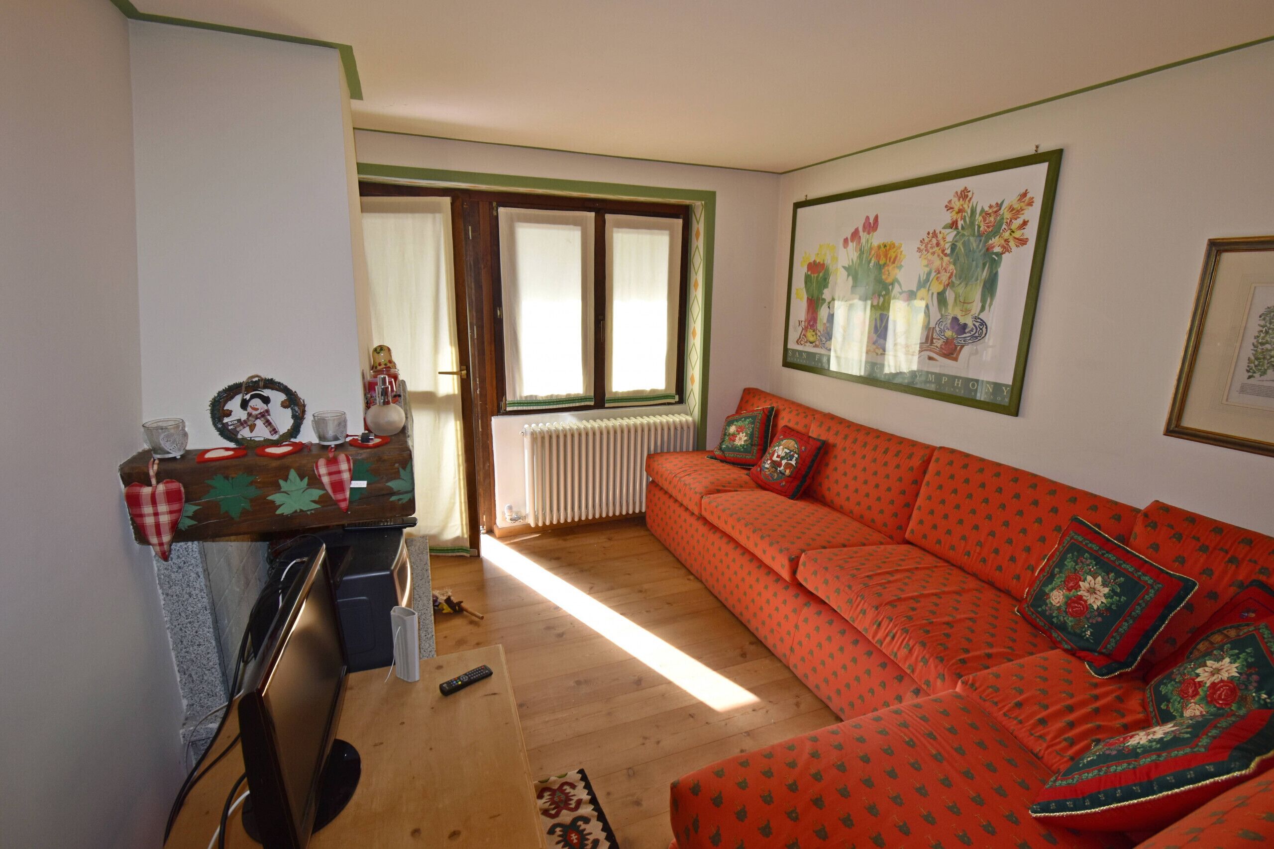 A living room with a wooden floor, a red sofa with patterned cushions, a wooden table with a heart-shaped decoration, and a framed picture on the wall.