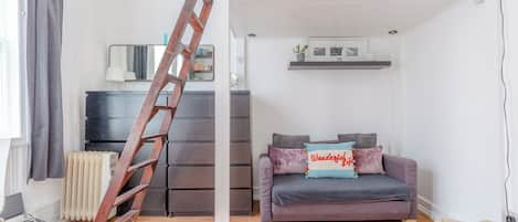 A modern living room with a wooden ladder, a grey sofa, and a small white shelf with decorative items.