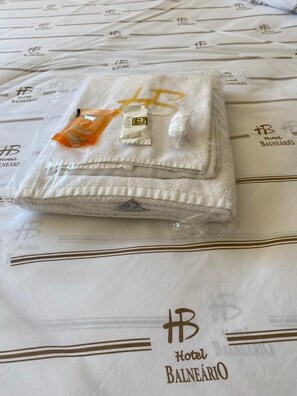 A neatly folded towel with a small orange packet on a Hotel Balneario branded bedspread. - Hotel Balneário (Poços de Caldas)