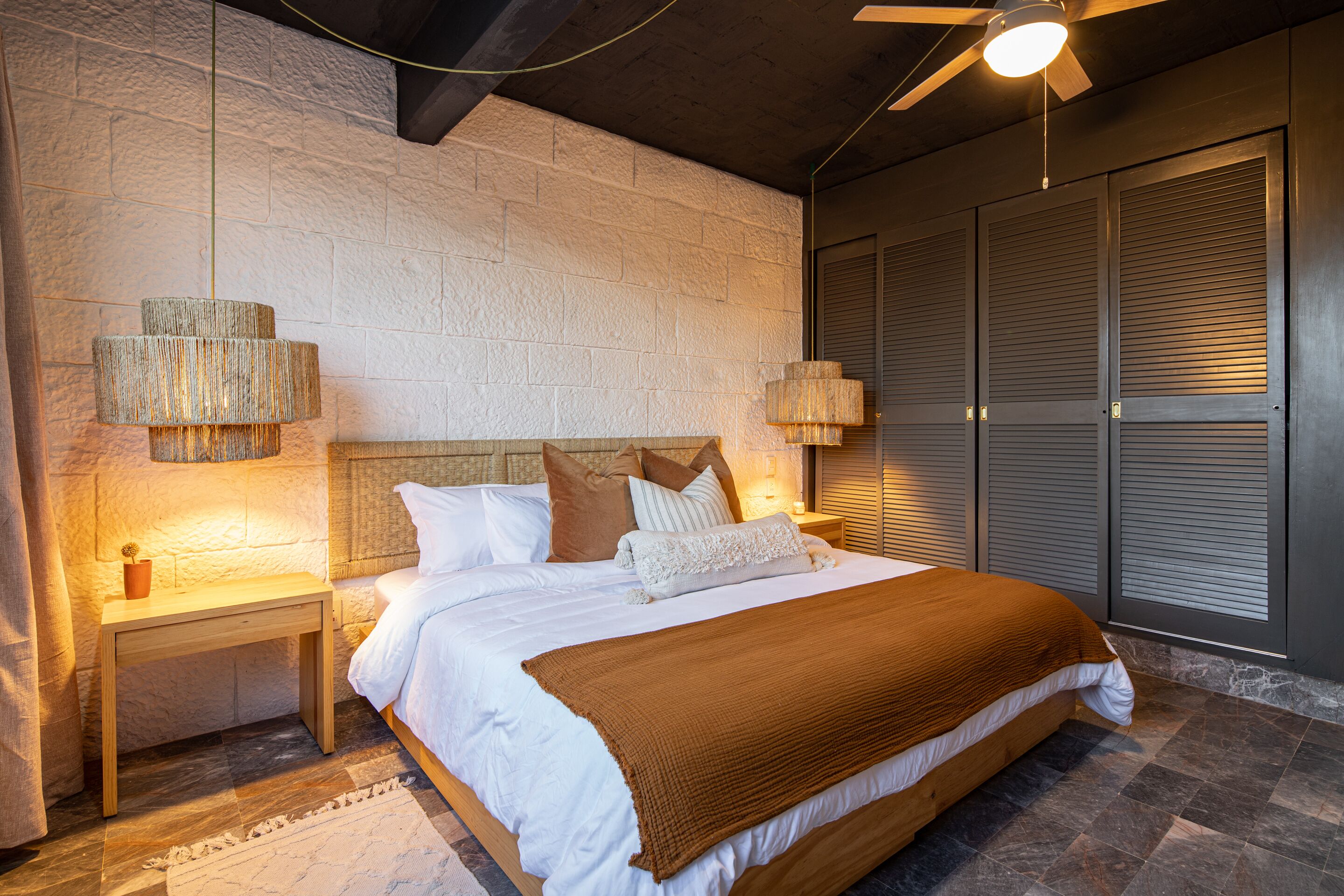 Ein Schlafzimmer mit einem großen Bett, zwei Nachttischen, einem Deckenventilator und einer Steinwand.