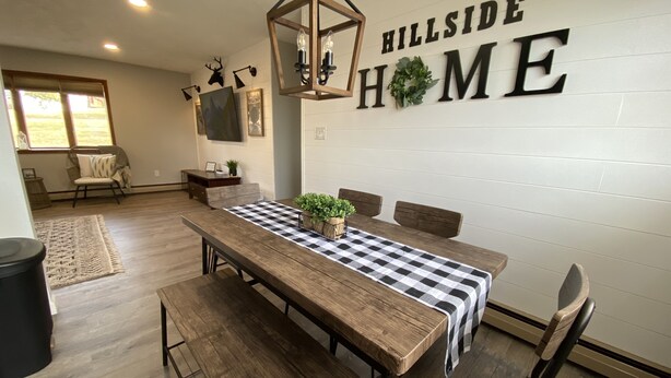 A dining area with a wooden table, checkered tablecloth, and a potted plant. A wall-mounted sign reads "HILLSIDE HOME".