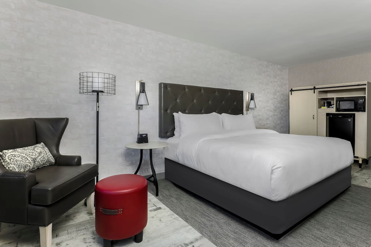 A modern hotel room with a large bed, a sofa, a red ottoman, a side table with a lamp, and a kitchenette.