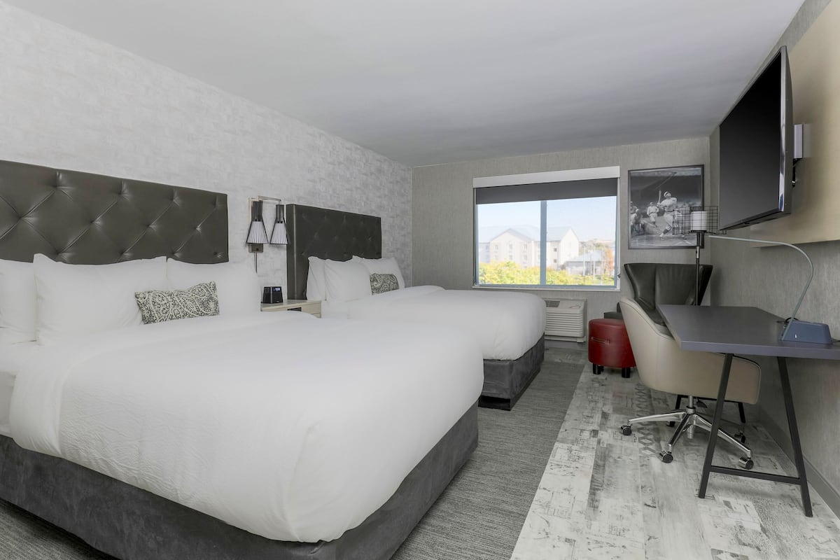 A hotel room with a large bed, a desk, a chair, a TV, and a window with a view of buildings.