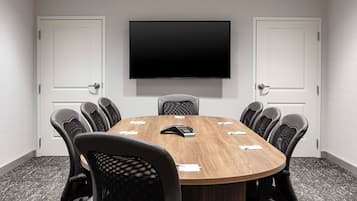 A conference room with a round table, six chairs, a flat-screen TV mounted on the wall, and two white doors.