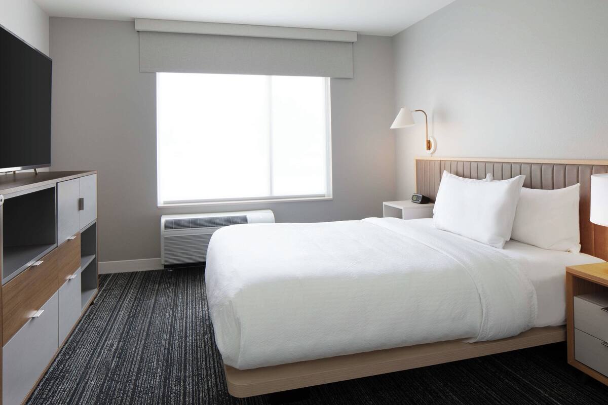 A hotel room with a bed, a TV, a bedside table, a lamp, and a window.
