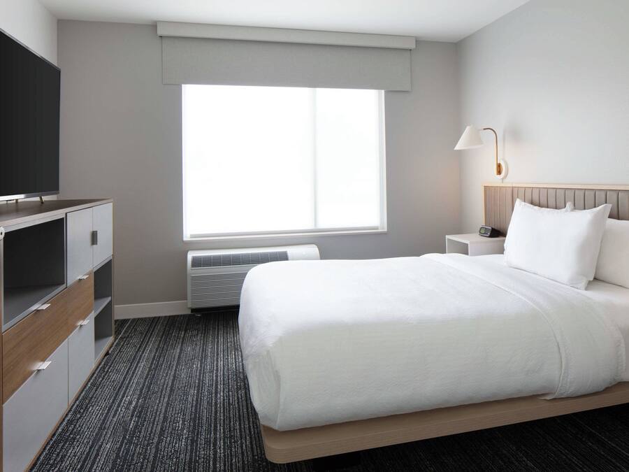 Una habitación de hotel con una cama, televisor, mesita de noche, lámpara y ventana.