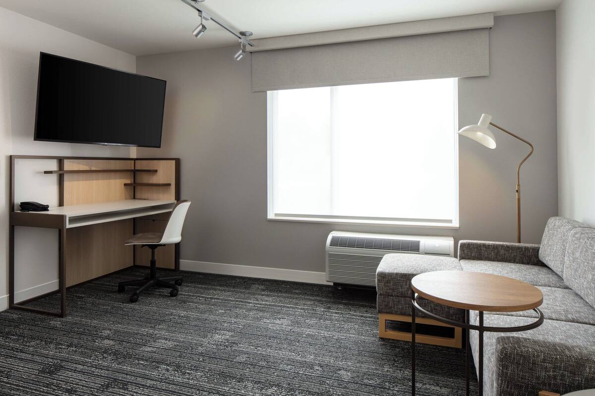 A modern room with a flat-screen TV, a desk with a chair, a sofa, a round table, and a standing lamp.