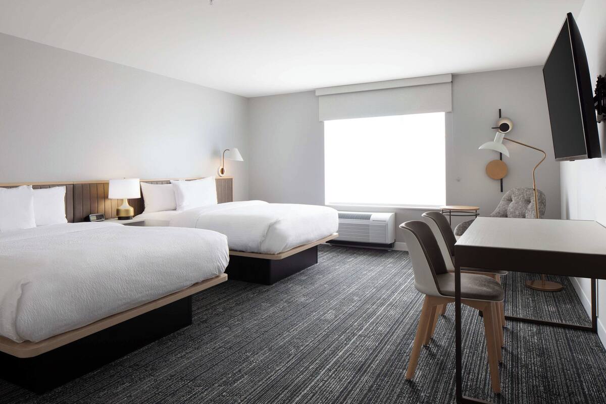 A hotel room with two beds, a desk, a chair, a TV, and a window.